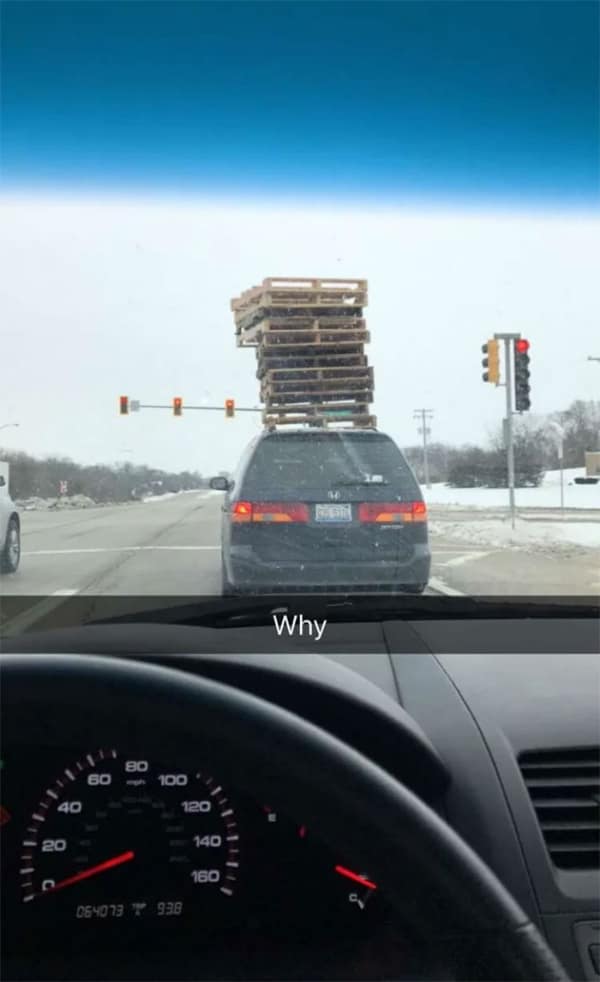 A dark minivan on a snowy road carrying an dangerously high, leaning stack of roughly fifteen wooden pallets on its roof. Text: "Why".