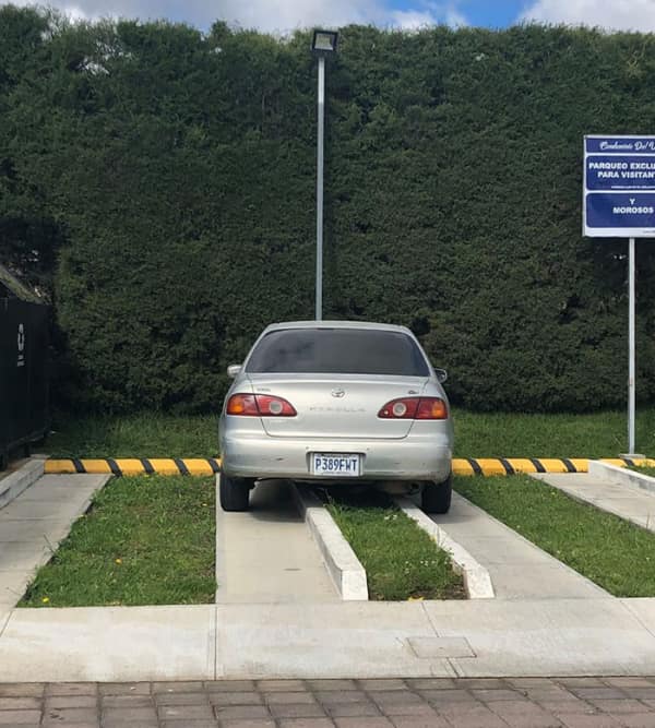 A silver sedan parked perfectly centered on a narrow concrete median strip meant to separate parking bays.