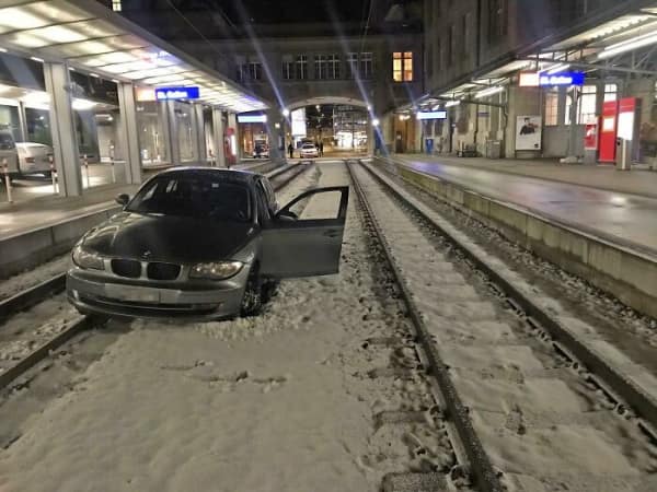car fail: A grey BMW sedan stranded on snow-covered train tracks at a transit station with the driver's side door open.