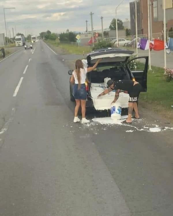 car fail: Two people on a roadside attempting to dump and clean a massive white paint spill from the interior of a car trunk