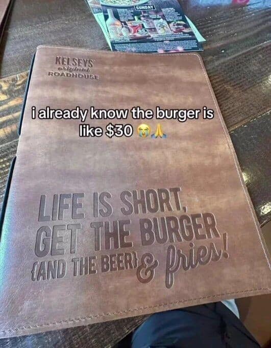 An all-too-real restaurant old meme showing a leather menu for Kelseys with the cheesy quote "Life is short, get the burger," paired with a customer's painful realization that the burger is definitely going to cost $30.