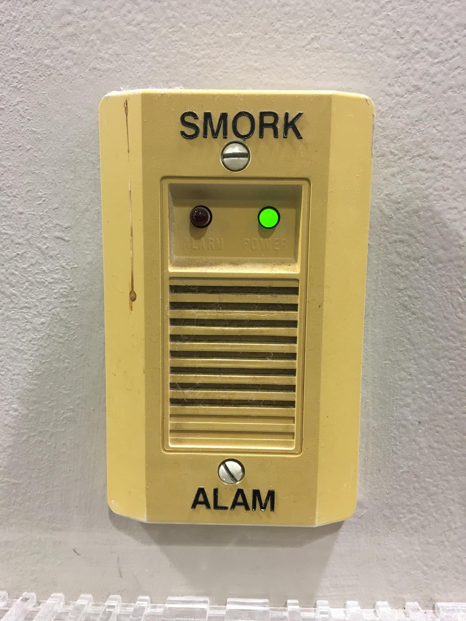 A phenomenal spelling fail captured in a classic meme, showing a standard beige wall-mounted fire detector that inexplicably and hilariously reads "SMORK ALAM."