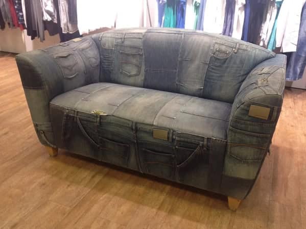 A weird furniture sofa upholstered entirely in patchwork denim jeans, featuring random pockets and waistbands scattered across the cushions.