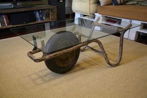 A weird furniture coffee table made by placing a glass sheet over a rusty, dirty wheelbarrow, bringing construction site chic to the living room.