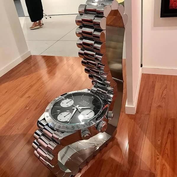 A weird furniture chair designed to look like a giant luxury wristwatch, where the metal link bracelet curves to form the seat and backrest.