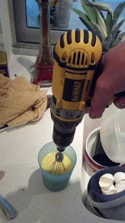 Hand holding a power drill with a whisk attachment to scramble eggs in a plastic cup.