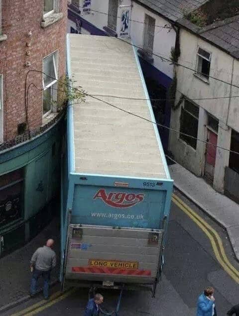 Large blue Argos delivery truck wedged tightly between two old brick buildings on a narrow street.