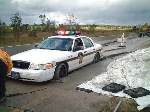 Police cruiser stuck deep in freshly poured wet cement on a highway construction site.