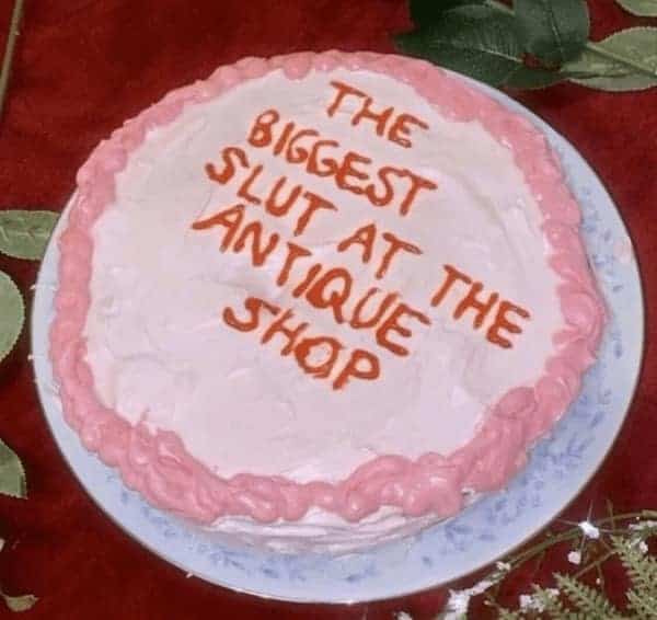 A pink frosted funny cake with a very specific insult declaring the recipient the biggest slut at the antique shop.