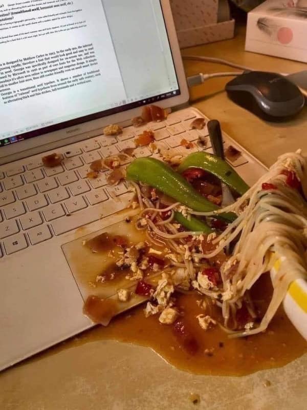 The ultimate home office funny fail showing a devastating spill where a massive bowl of soupy noodles and vegetables is dumped directly across the keyboard of an expensive white laptop.