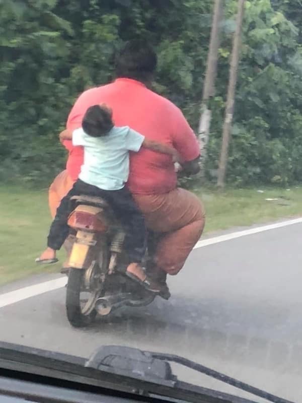 A chaotic transportation bad day photo showing a massive man driving a motorcycle on a rural road while a small child sits entirely backwards behind him, completely passed out and slumped over his back.
