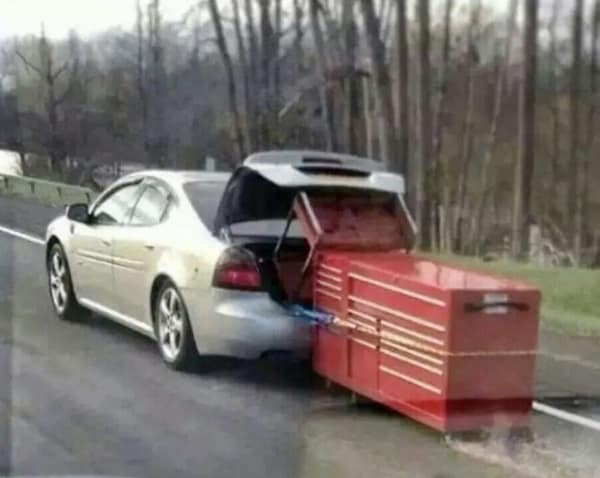 A redneck engineering funny fail featuring a silver sedan driving down a rural road while towing a massive red mechanic's tool chest directly by its handle on tiny caster wheels.