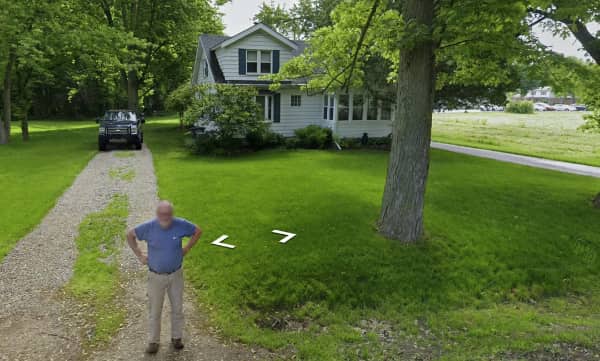 An elderly man standing on a gravel driveway looking confused at the Google Street View car.