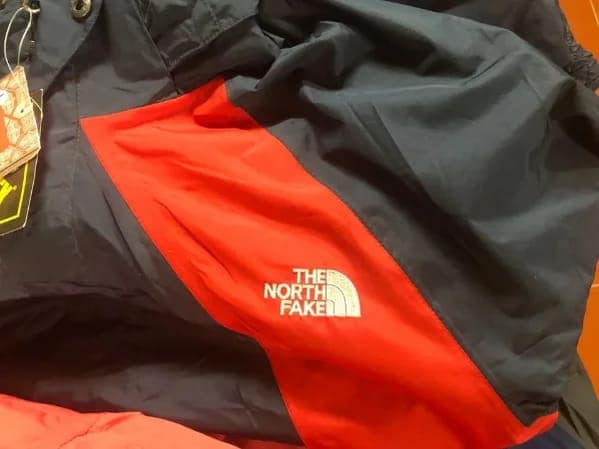 Red and black winter jacket featuring an embroidered logo reading The North Fake. Self aware bootleg outdoor fashion.
