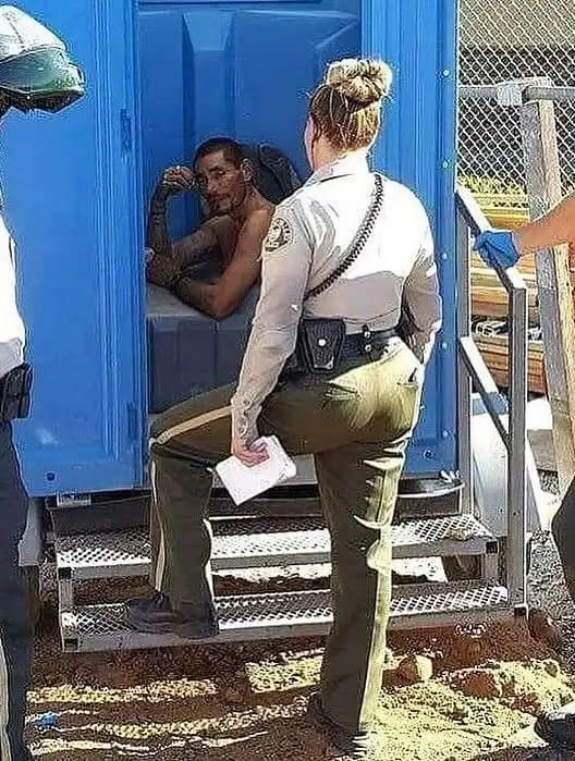 Female police officer standing in front of a portable toilet with a man sitting inside.
