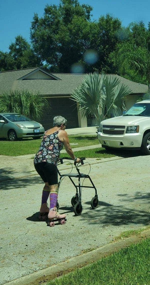 Elderly woman using a medical walker for stability while wearing pink roller skates on a residential street.