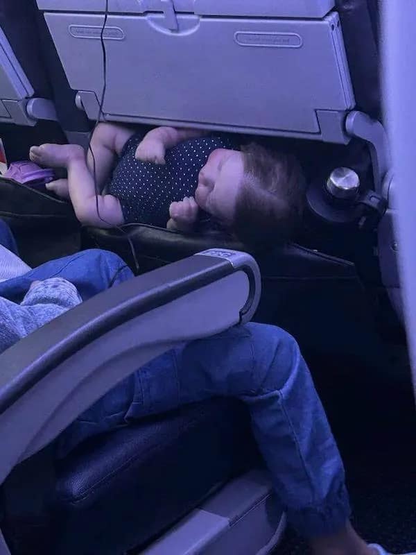 A hilarious meme capturing a baby sleeping comfortably tucked inside the seatback pocket of an airplane seat like a kangaroo pouch.