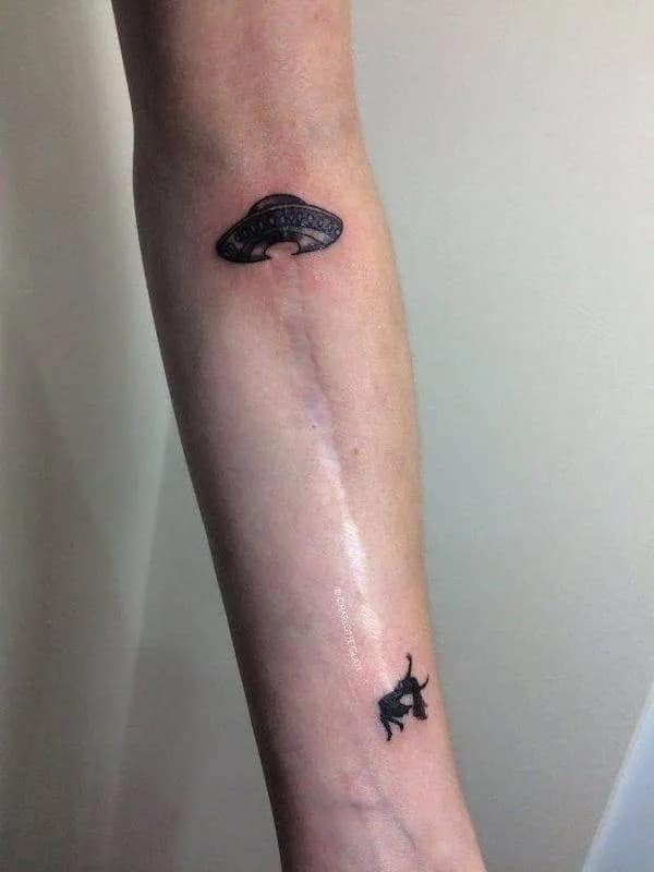 A creative hilarious meme tattoo where a long surgical scar is turned into an alien abduction beam lifting a stick figure to a UFO.