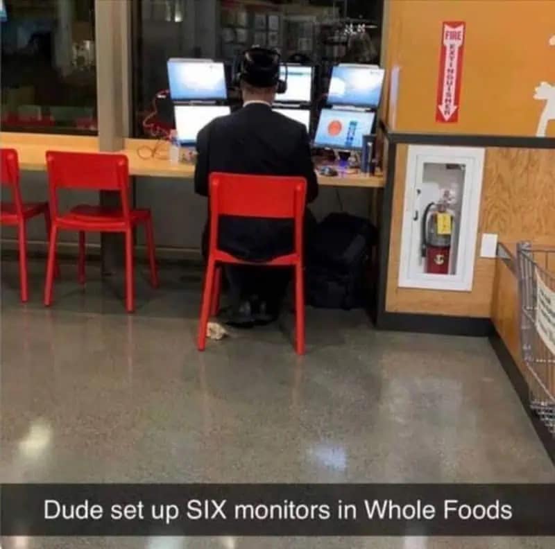 A wildly chaotic meme dump image capturing a man in a dark suit sitting at a grocery store counter who has completely taken over the space by setting up an absurd workstation with six different computer monitors, captioned "Dude set up SIX monitors in Whole Foods" in this funny meme.