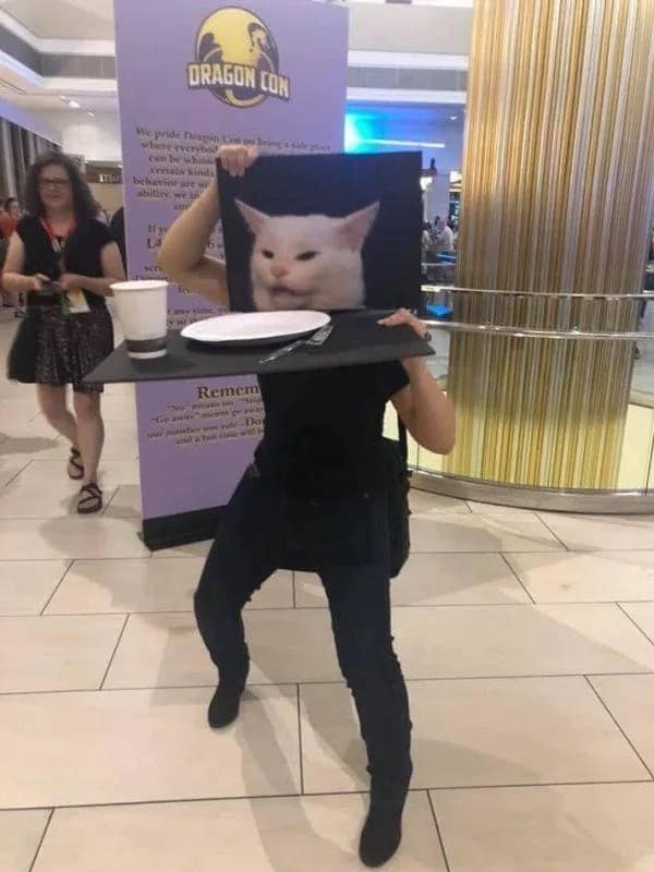 Dragon Con cosplayer wearing a mask of Smudge the white cat sitting behind a dinner plate from the famous yelling meme.