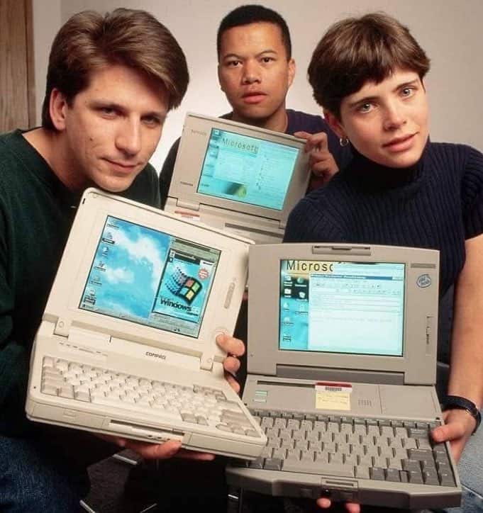 Three people posing while holding vintage 1990s laptops with thick bezels and trackballs.