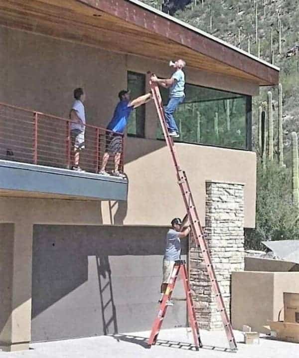 A terrifying osha meme showing three men creating a "ladder tower" by stacking a step ladder on top of an extension ladder to reach a roof, while the guy on top casually takes a drink.