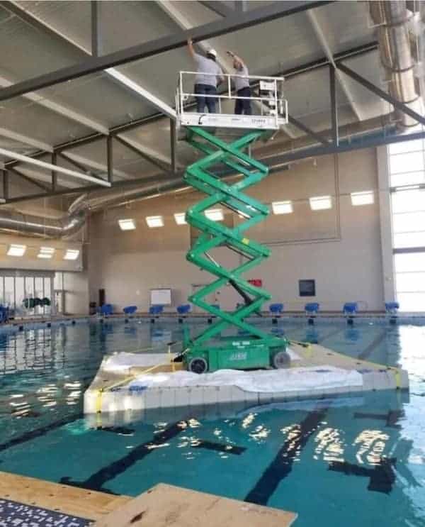 A mind-blowing osha meme featuring a heavy scissor lift placed on a floating raft in the middle of a swimming pool so workers can reach the ceiling lights.