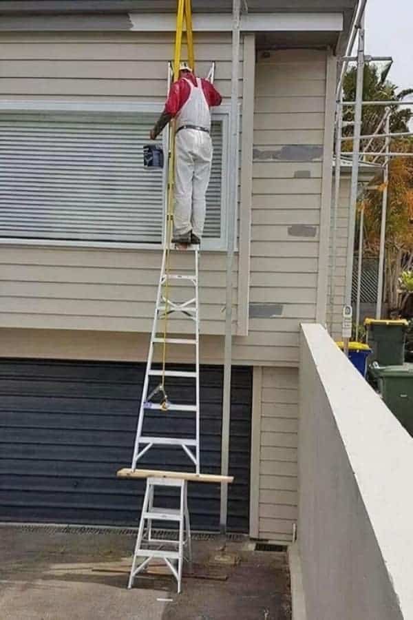 A creative but dangerous funny osha fail showing a painter balancing a tall extension ladder on top of a small step ladder to reach a second-story window.