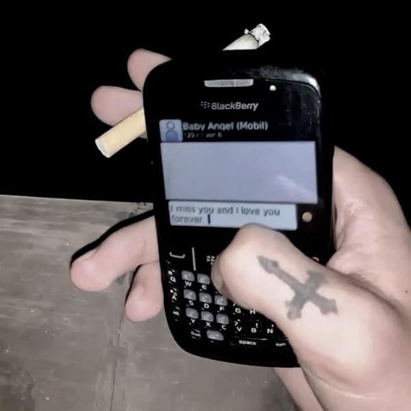 A "toxic ex" style relationship meme aesthetic featuring a hand with a cross tattoo holding a cigarette and a BlackBerry, sending a desperate "I miss you" text.