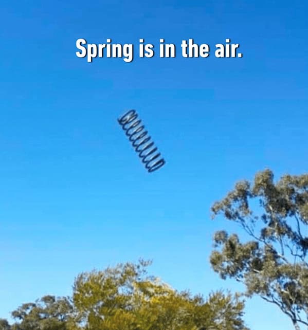 A brilliantly literal spring meme showing a large metal coil bouncing high into the bright blue sky above some trees, accompanied by the classic seasonal pun, "Spring is in the air".