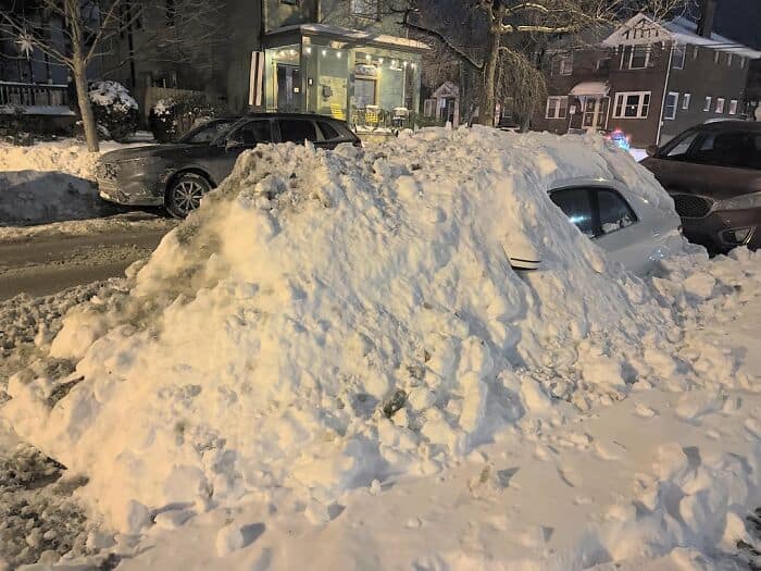 White car completely buried under a massive mountain of snow as winter parking revenge.