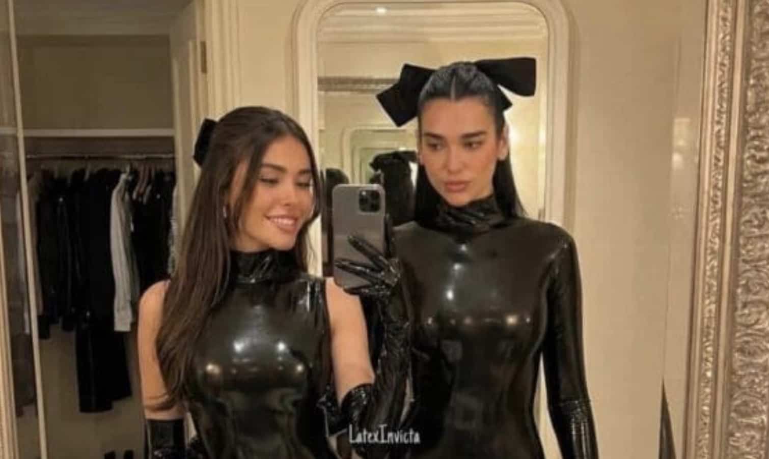 Alt Text: A mirror selfie of two women, Madison Beer and Dua Lipa, posing in a dressing room. They are both wearing matching shiny black latex outfits—one a sleeveless turtleneck and the other a long-sleeved turtleneck—complete with large black bows in their hair and matching latex gloves.