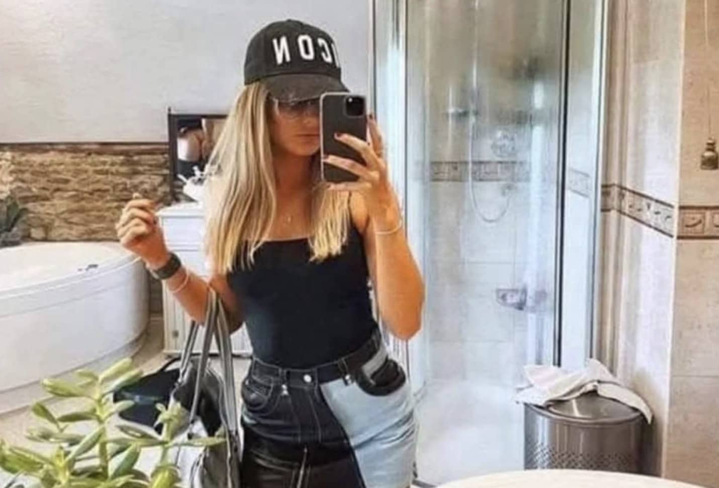 A woman taking a mirror selfie in a bathroom with an accidental reflection visible in the background.