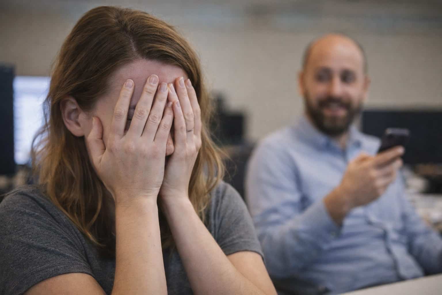 Woman covering her face in embarrassment while a man in the background smiles at his phone.