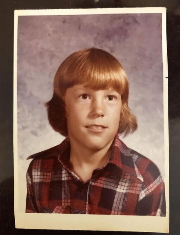 A young boy in a plaid shirt wearing a quintessential 80s haircut "mushroom" cut. The look features a massive volume of straight hair on top that curves sharply inward at the ears, creating a heavy, rounded silhouette.