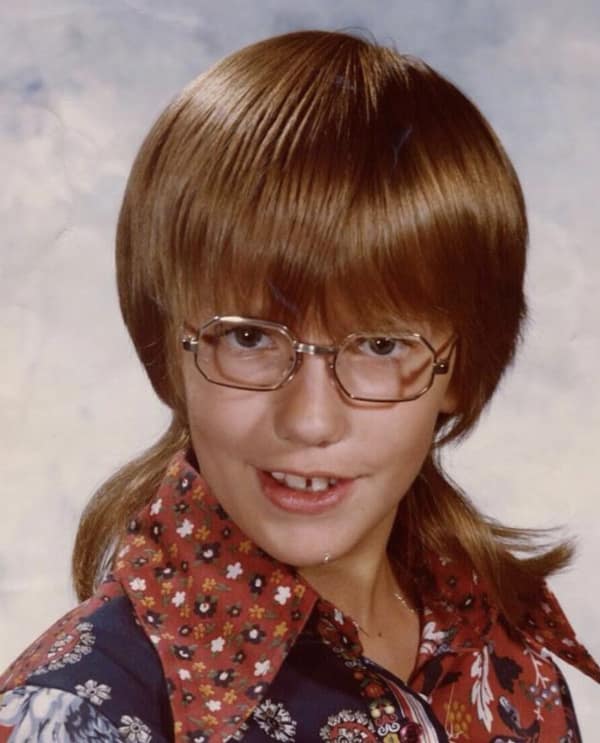80s haircut mullet-shag hybrid on a young boy wearing large glasses. The hair is thick and rounded on the top and sides but falls into thin, shoulder-length layers in the back.