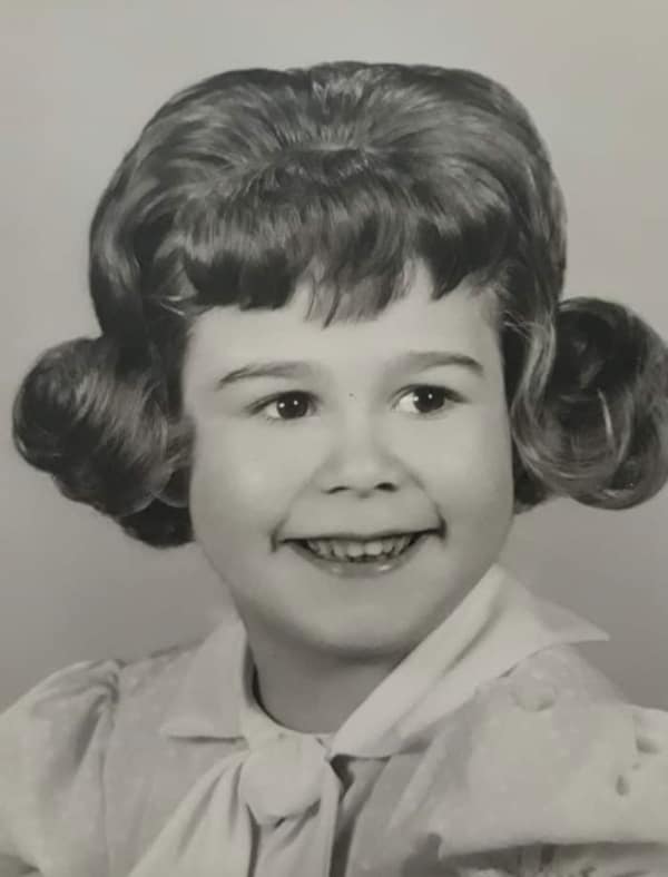 A black-and-white portrait of a young girl with a gravity-defying 80s haircut. The style features a massive, hairsprayed "pouf" on top and hair curled into large, round loops at the sides, representing extreme formal styling of the era.