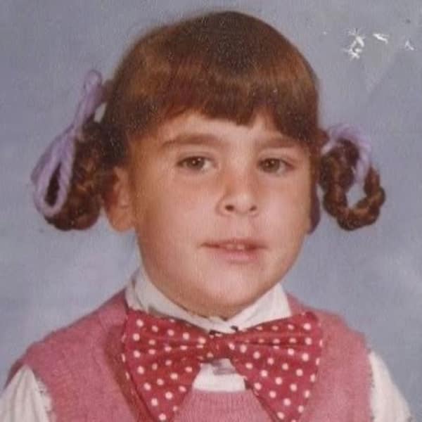 young girl with a highly structured 80s haircut featuring thick, blunt bangs and pigtails that have been tightly braided and looped back up into "buns" with purple ribbons.