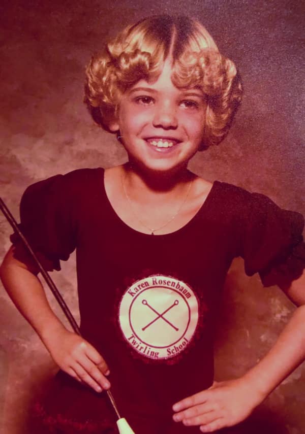 A smiling young girl in a "Twirling School" leotard wearing an iconic 80s haircut composed of tight, structured ringlet curls. The perm-heavy style frames her face in a halo of bouncy, golden spirals.