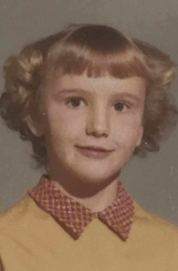 young girl with a structured, blonde 80s haircut. The style features short, straight bangs paired with hair curled outward into distinct "wings" at the sides, a very popular look for school-aged children in the early 80s.