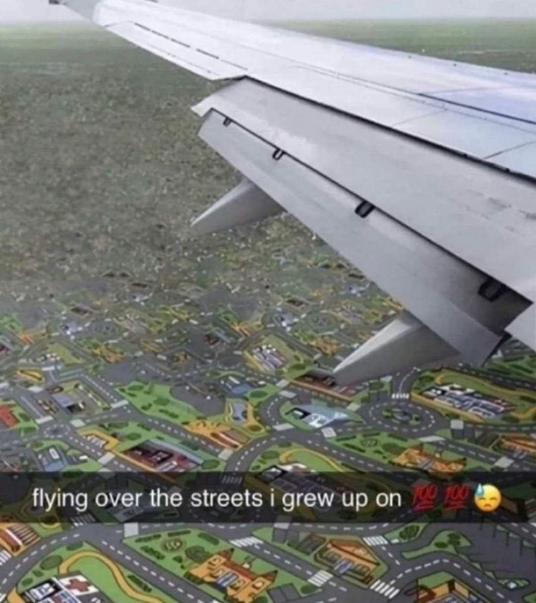 clever 90s meme showing an aerial airplane wing view over a landscape that is actually a classic childhood play rug featuring roads and cartoon buildings, captioned "flying over the streets i grew up on."