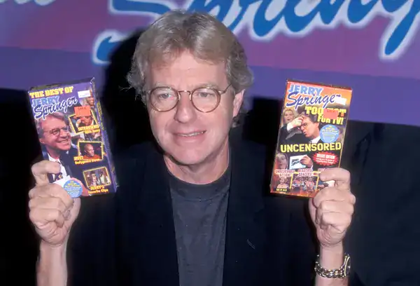 legendary daytime television moment in 90s pictures featuring host Jerry Springer smiling while holding up two VHS tapes of his own show, including "The Best of Jerry Springer" and "Too Hot for TV".