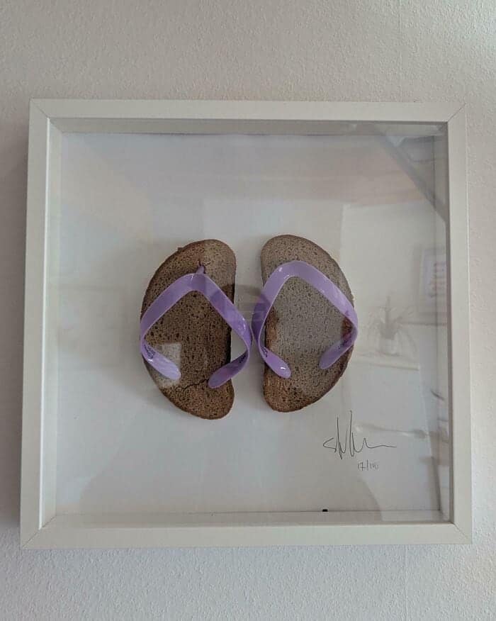 Two slices of brown bread fitted with purple plastic straps to look like flip-flop sandals framed as modern art. An absurd diy fail.