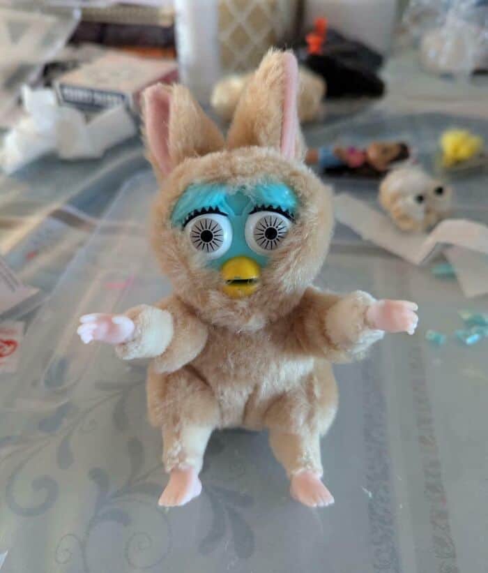 A deeply cursed toy featuring a Furby face attached to a fluffy bunny body with realistic human baby hands and feet. A nightmare hilariously bad DIY.