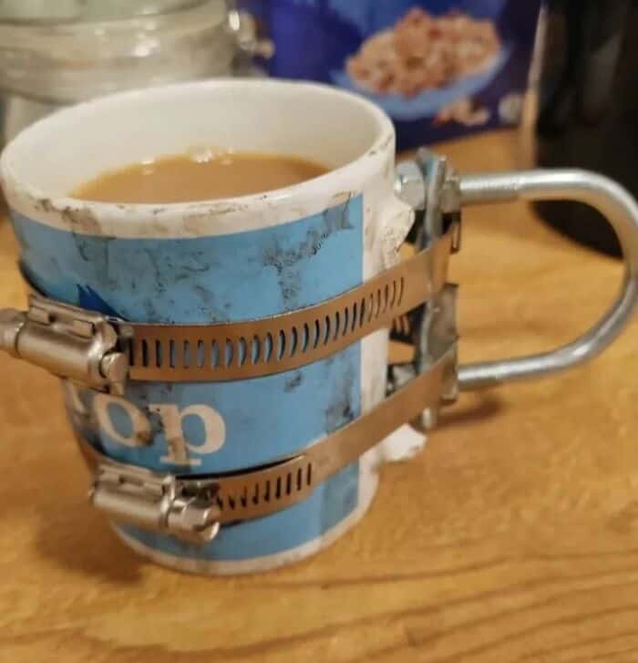 A broken ceramic coffee mug brilliantly fixed by strapping a heavy metal U-bolt handle to the side using two industrial hose clamps in this hilariously bad DIY.