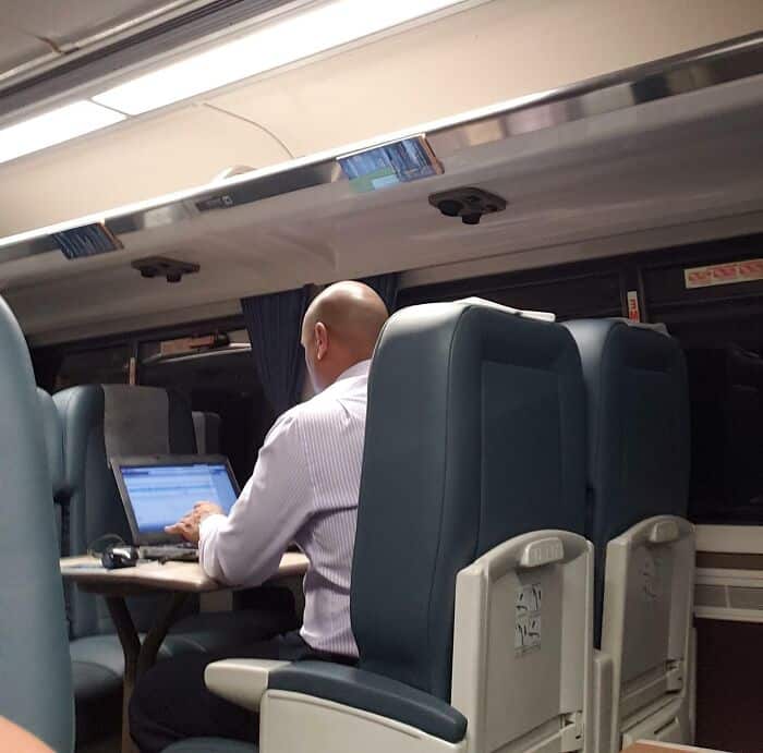 passenger turns a shared train car into a private cubicle, hunched over his laptop and dominating the table space in a common struggle with annoying people on public transit