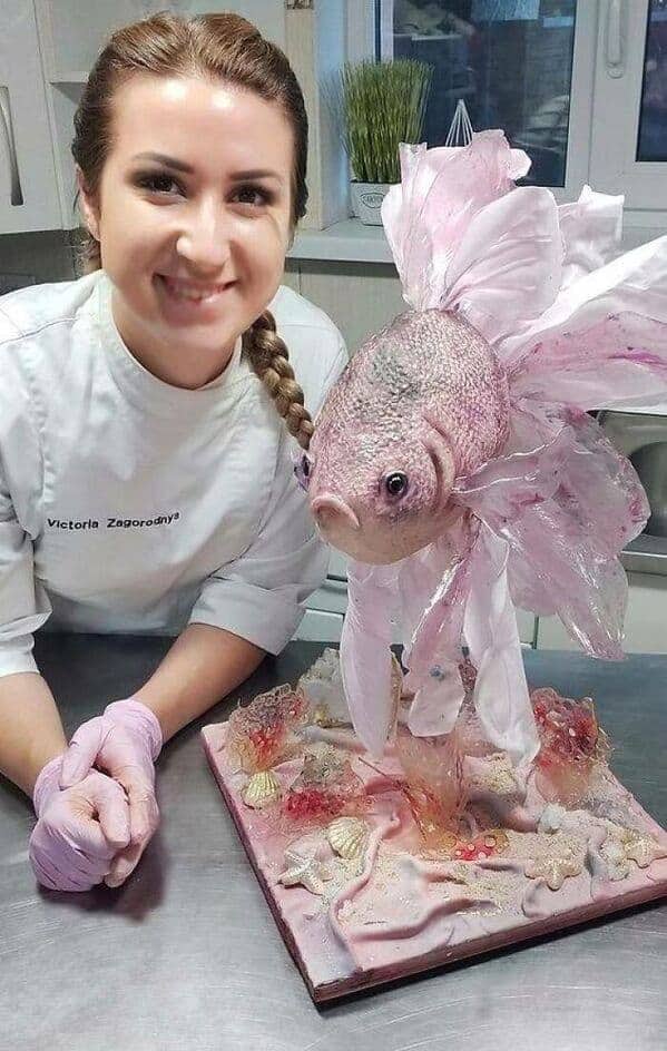 A professional pastry chef posing with an incredibly realistic pink goldfish cake on a coral base.