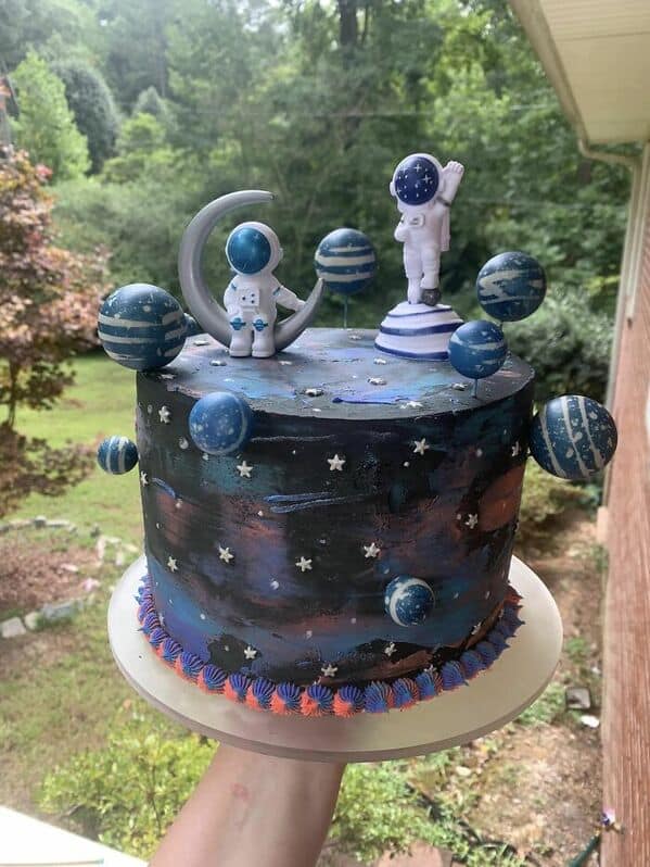 A dark blue outer space cake decorated with astronauts, planets, and a crescent moon topper.