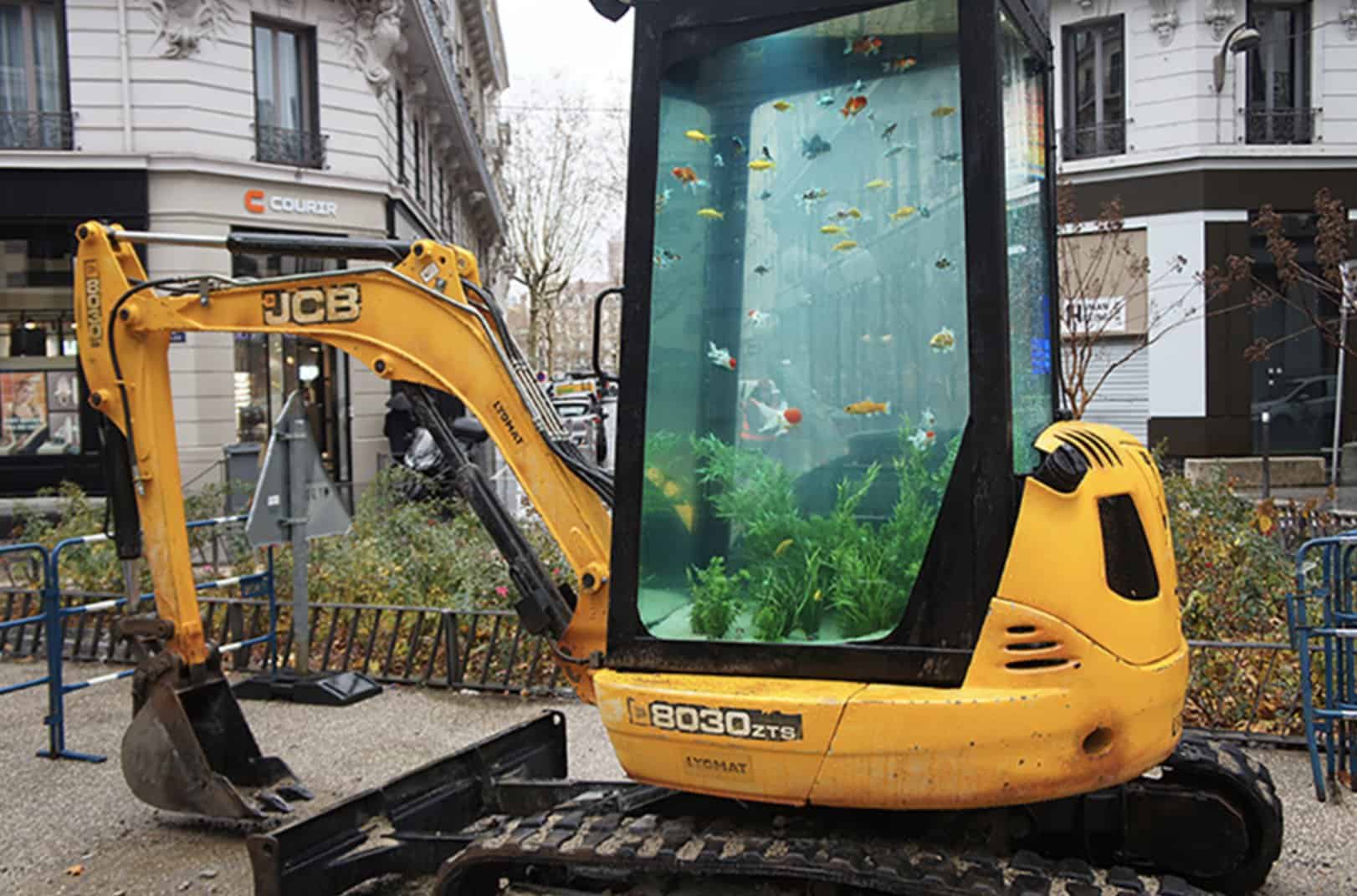 Street art installation featuring a small yellow excavator with a fish tank cab.