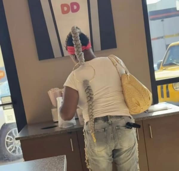 An absolutely unhinged bad haircut captured inside a Dunkin' Donuts, showing a woman with a long blonde braided extension violently looping through a giant gold hoop earring before continuing down her back.